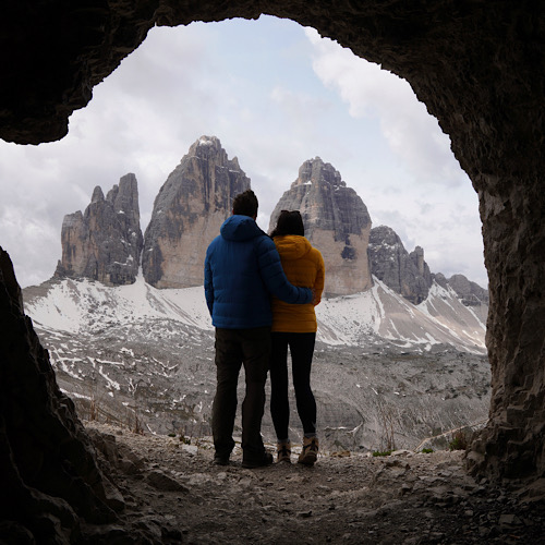 A couple sitting inside a mountain cave looking out at the Tre Cime di Lavaredo peaks.