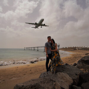Travel couple standing on a beach in Lanzarote directly under a low-landing airplane.