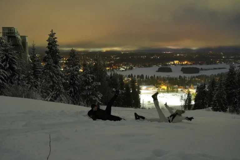 A couple lies in the snow on Ounasvaara hill in the evening, looking at the lights of Rovaniemi.