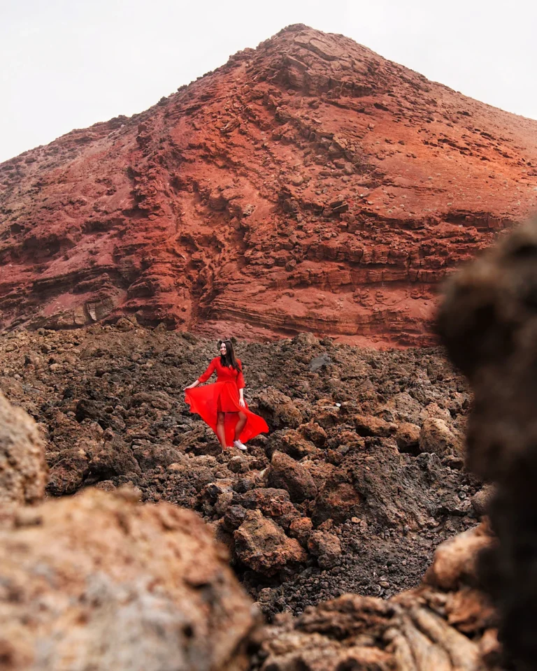 Frau im roten Kleid vor dem markanten roten Vulkan Montaña Bermeja auf Lanzarote.