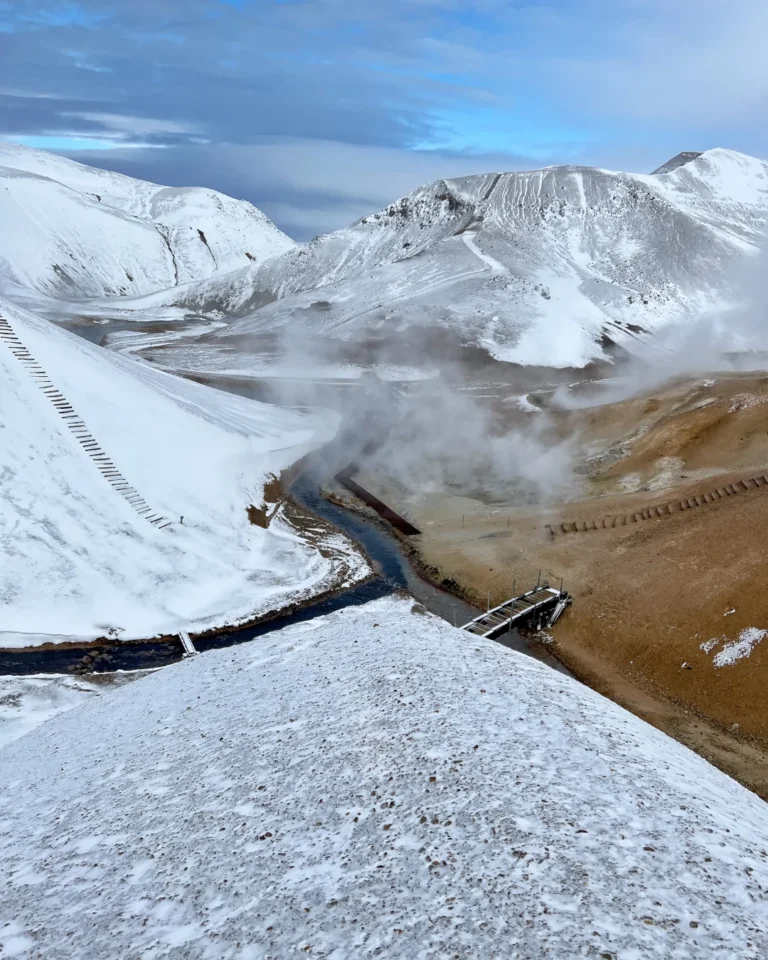 A scenic view of Hveradalir with geothermal steam, snow-capped mountains, and a pedestrian bridge, Kerlingarfjöll, Iceland