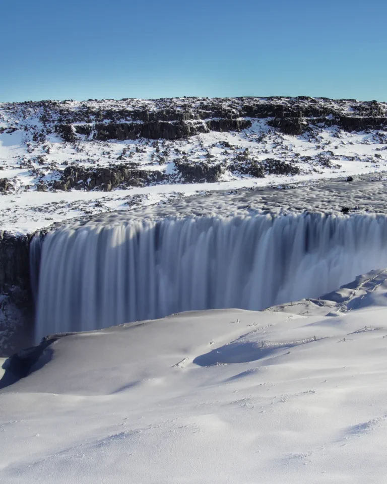 Dettifoss waterfall surrounded by snow and ice, with a clear blue sky above, Iceland