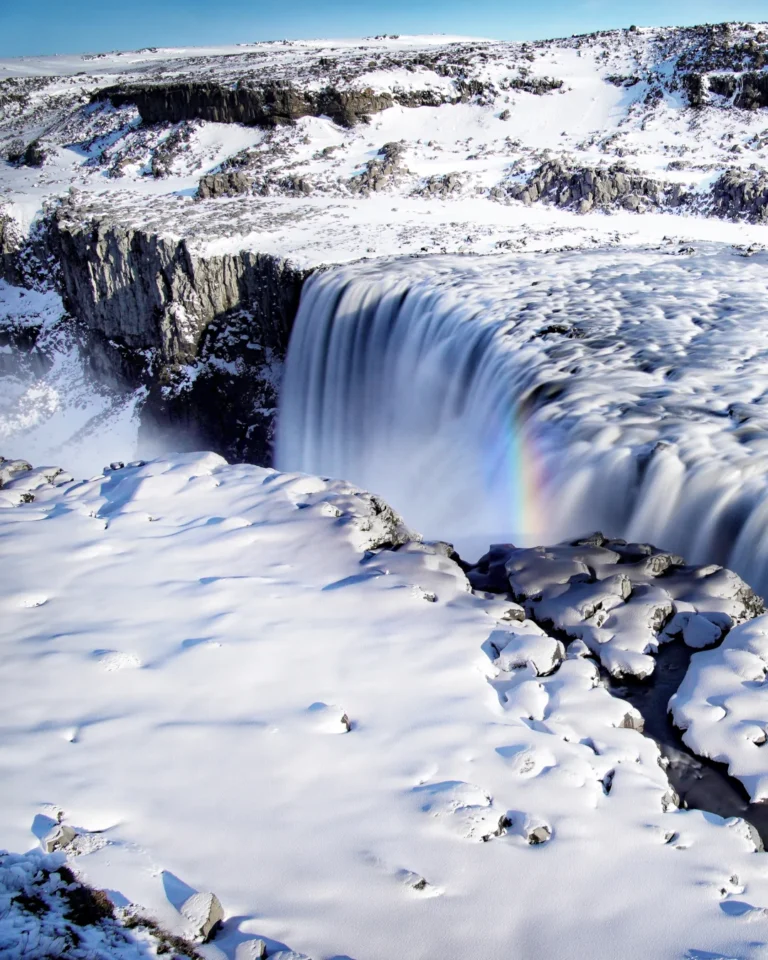 Dettifoss waterfall surrounded by snow and ice, with water flowing down and a little rainbow under a blue sky