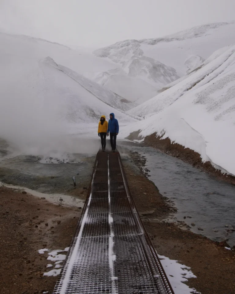 Two people standing amidst geothermal steam on a wooden bridge in Hveradalir, Kerlingarfjöll, Iceland