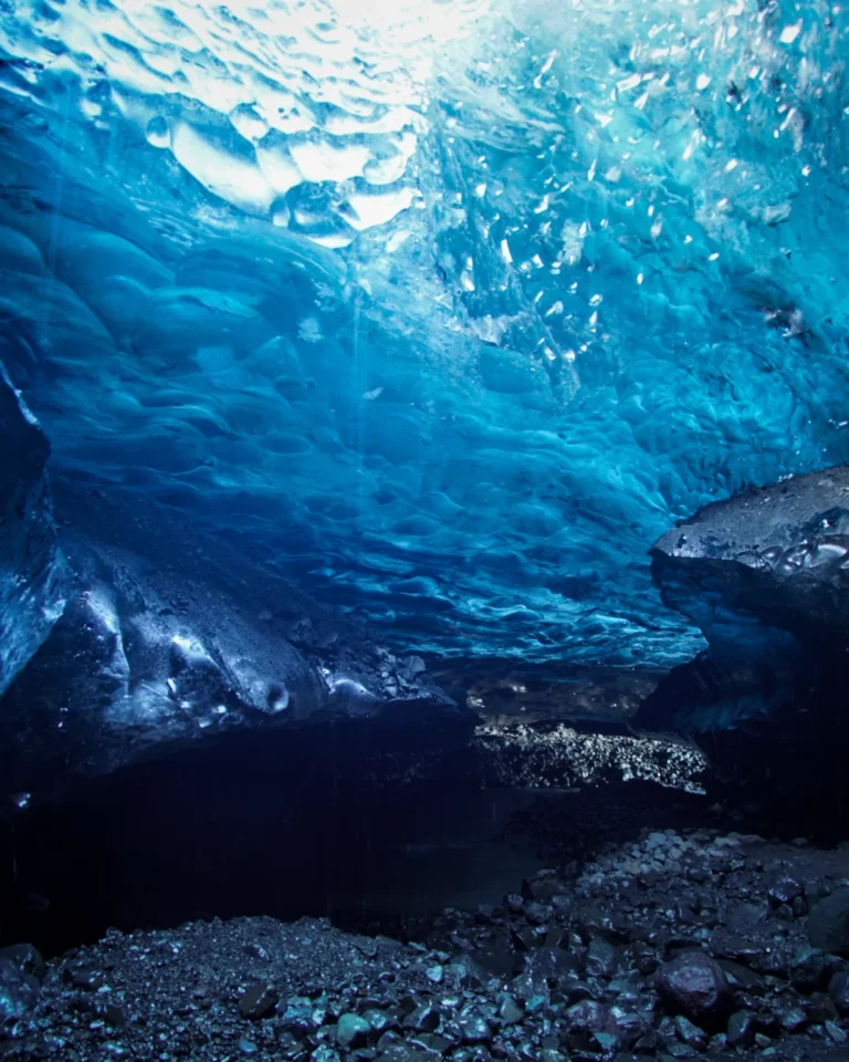 Inside a Vatnajökull ice cave, with shimmering blue light illuminating the ice-covered roof and a small river flowing through, Iceland