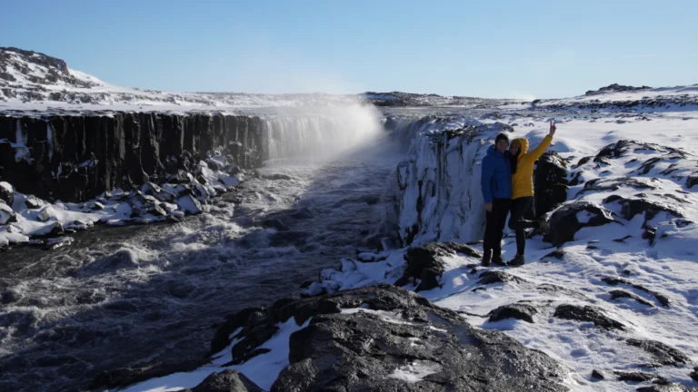 A couple standing in front of Selfoss waterfall, Iceland, with a clear blue sky above