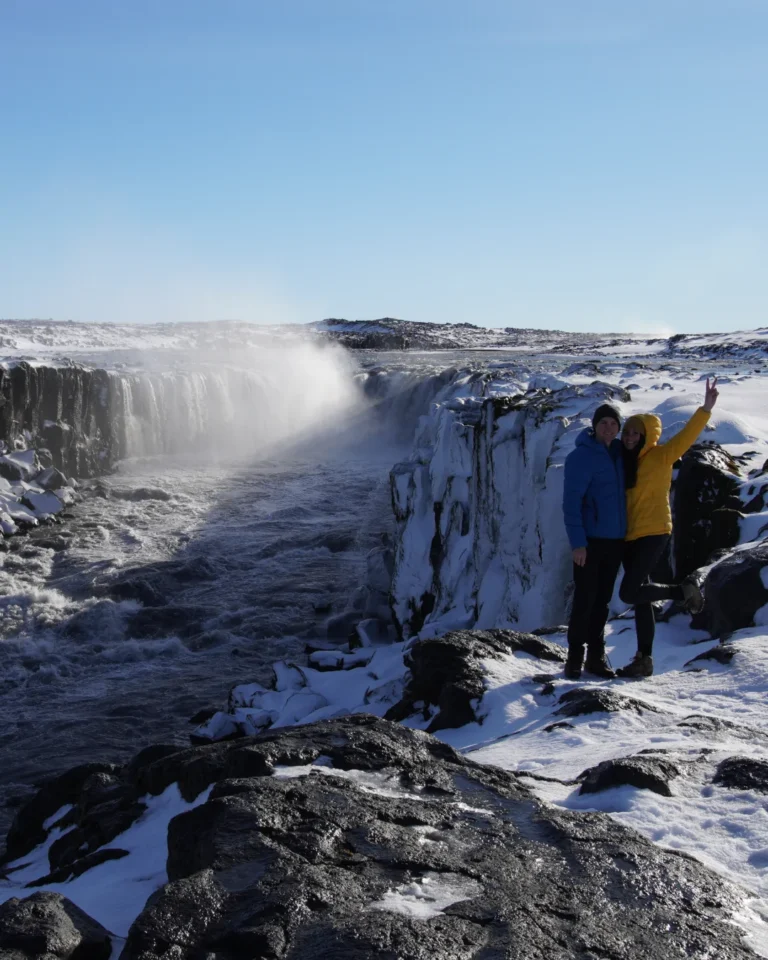 A couple standing in front of Selfoss waterfall, Iceland, with a clear blue sky above