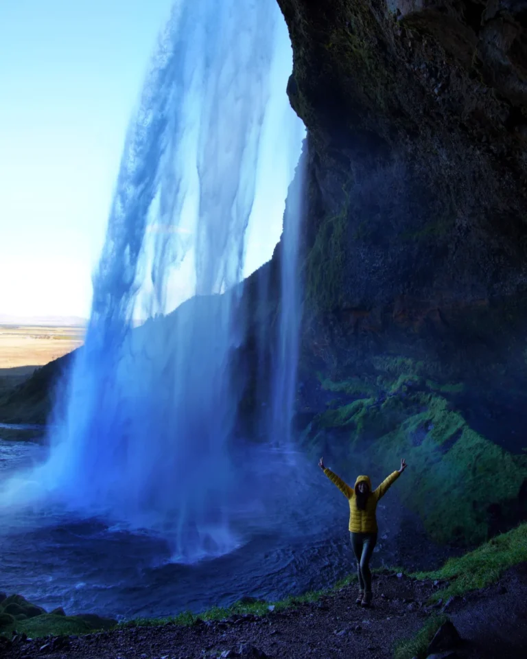 raising the arms in celebration behind Seljalandsfoss, with water falling and Icelandic greenery visible beyond