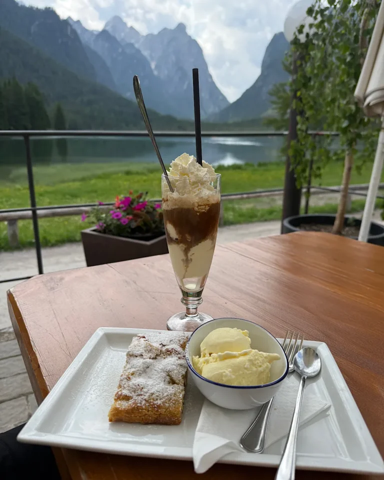 Ein Teller mit Apfelstrudel und Vanilleeis sowie ein Eiskaffee auf einem Holztisch vor einer Bergkulisse.