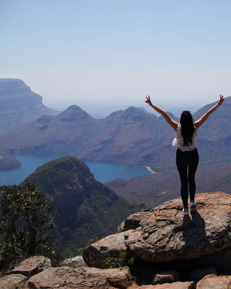 A woman standing with her arms raised at a viewpoint overlooking the Blyde River Canyon.