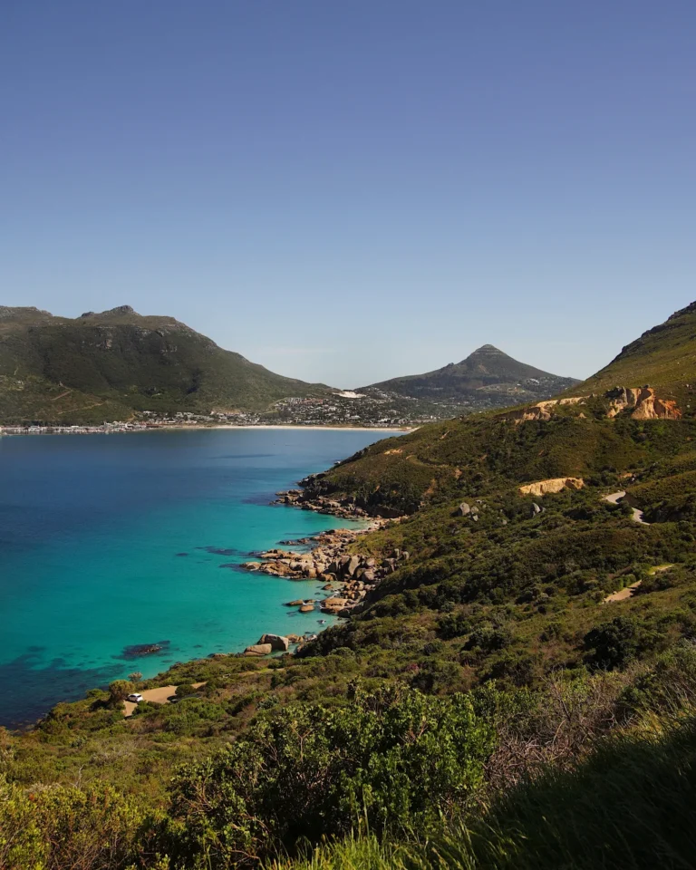 Winding Chapmans Peak Drive along green mountains with blue ocean and Lions Head in the background.