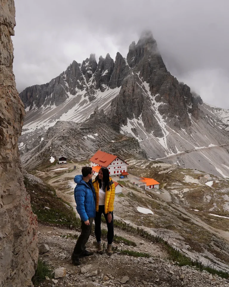 A couple standing on a trail in front of the red-roofed Locatelli Hut with the Paternkofel mountain in the background.