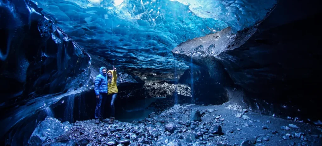 Paar in gelber und blauer Jacke unter der glänzenden blauen Eisdecke einer Gletscherhöhle am Vatnajökull in Island.