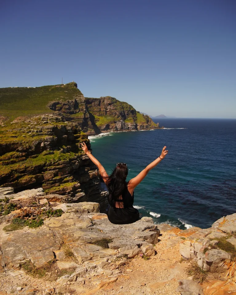 Back view of a woman sitting on a cliff at Cape of Good Hope with arms raised overlooking the blue ocean.