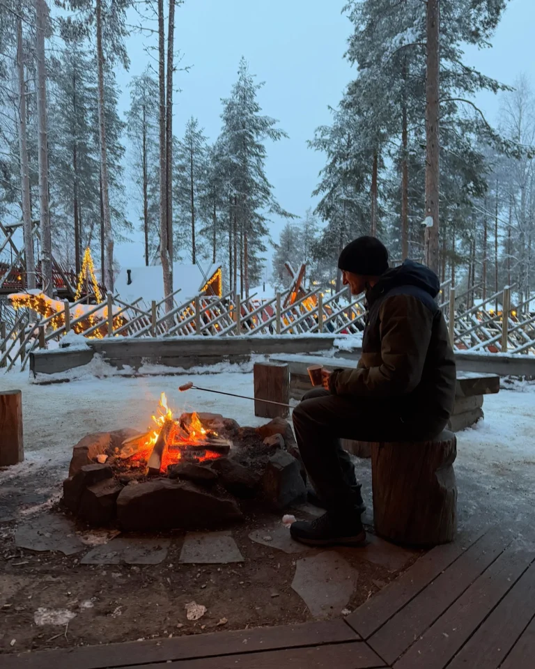 A man sitting by a campfire in Santa Claus Village Rovaniemi with mulled wine and a grill.