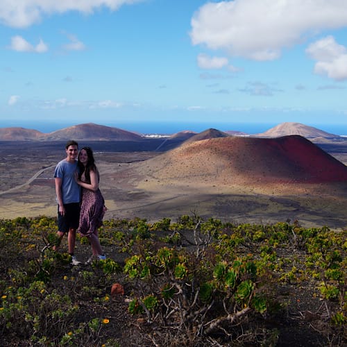 Couple standing on top of a volcano in Lanzarote overlooking a barren landscape and deep red volcanic cones.
