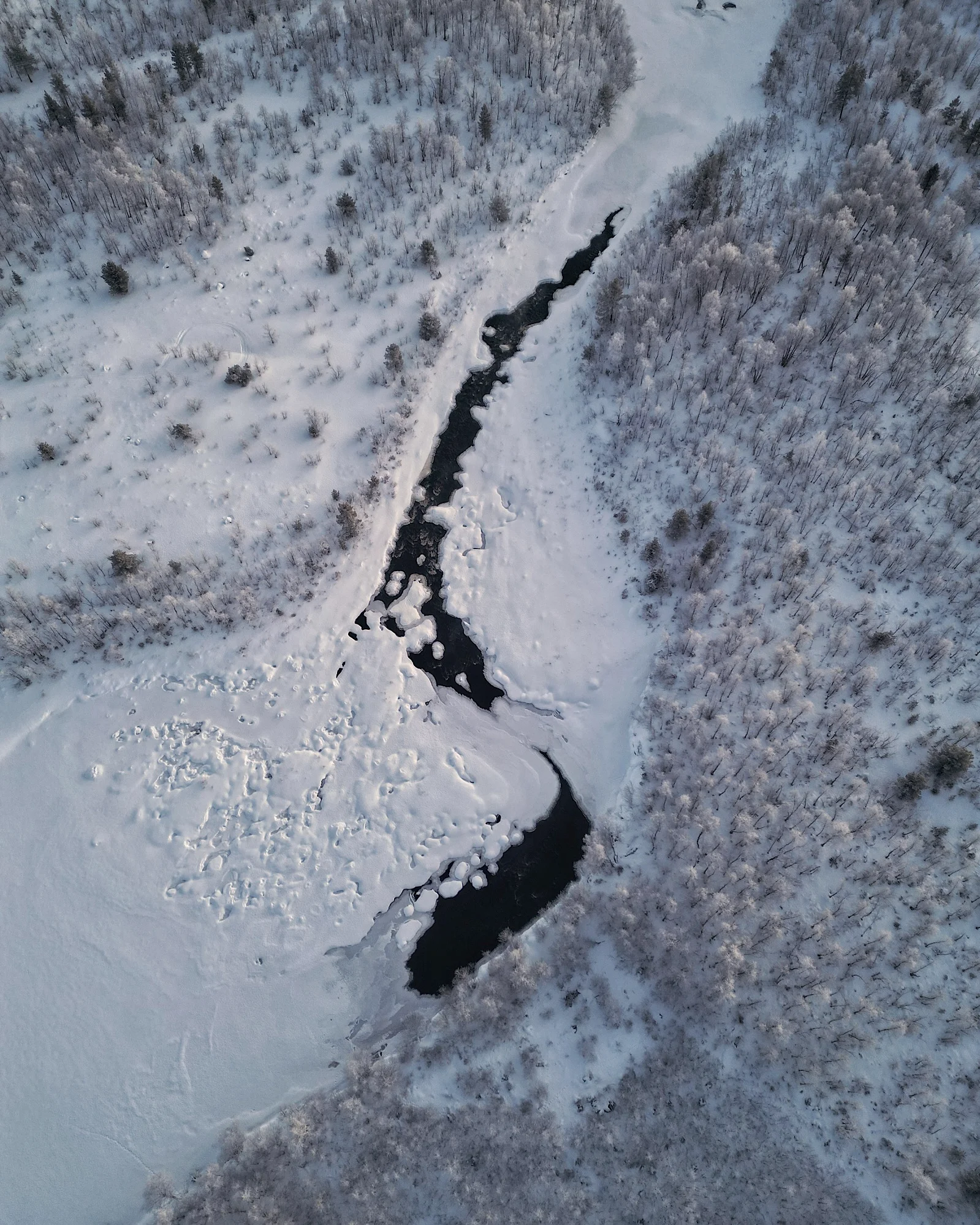 Drohnenfoto eines winterlichen Flusses in Lappland mit teilweise offenem Wasser und verschneiten Ufern.