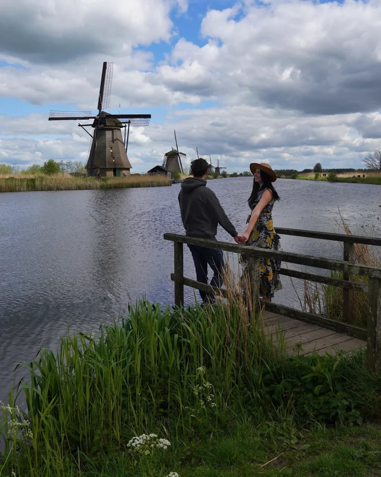 Ein Paar steht am Ufer in Kinderdijk und schaut sich vor der Kulisse eines Stegs am Wasser die Windmühlen an.