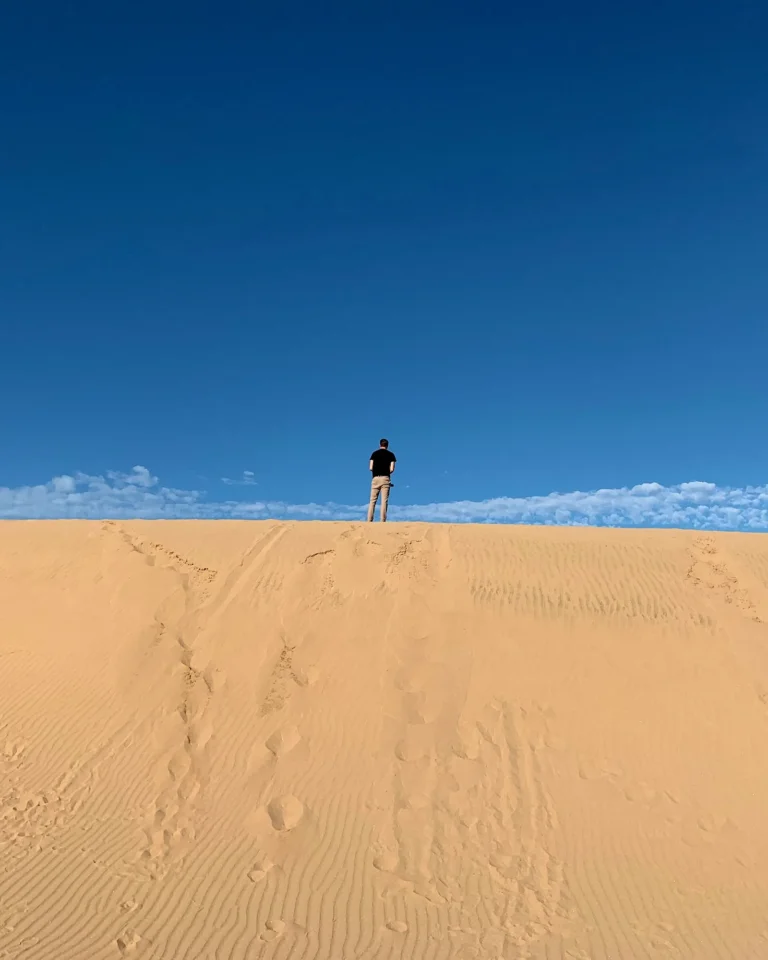 Man standing on top of a sand dune summit against the blue sky.