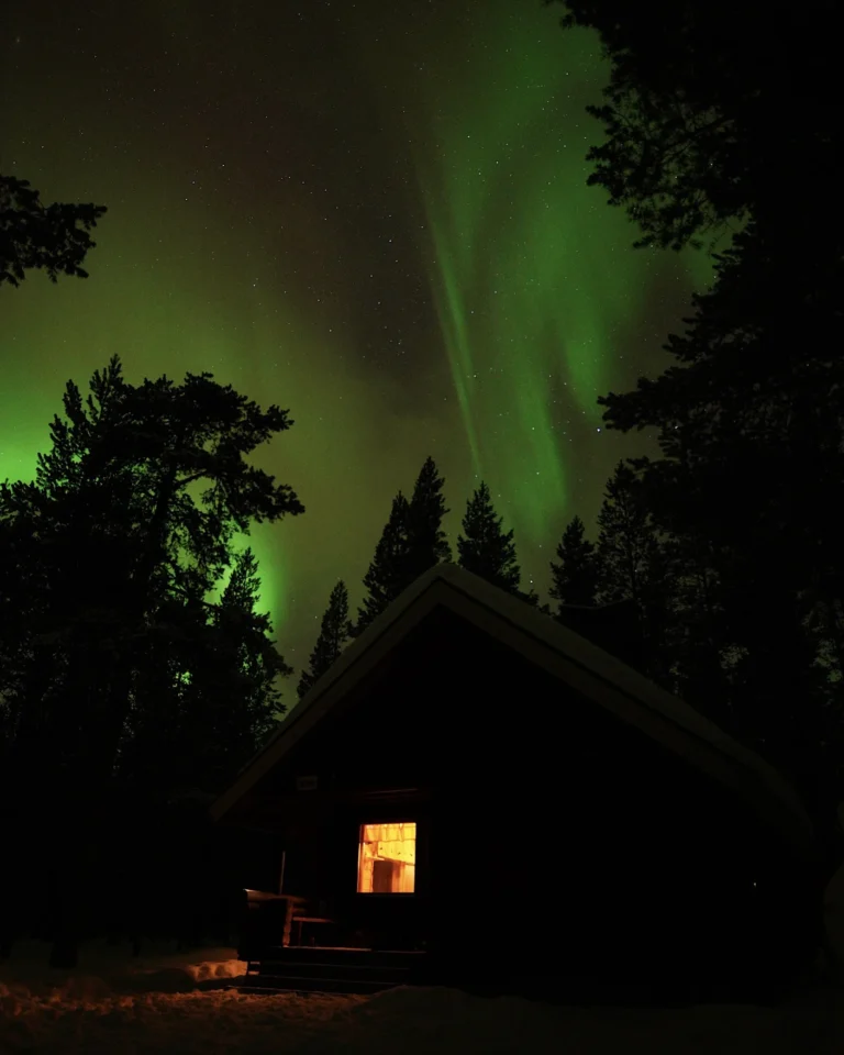 Vibrant green Northern Lights (Aurora Borealis) dancing in the night sky above a lonely, snow-covered wooden cabin in the Lapland wilderness.