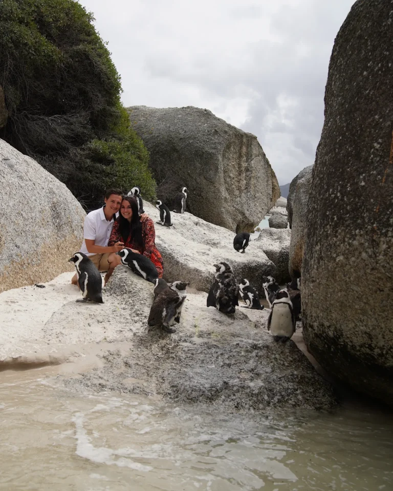 Travel couple looking into the camera at Boulders Beach with penguins on rocks and in the water.