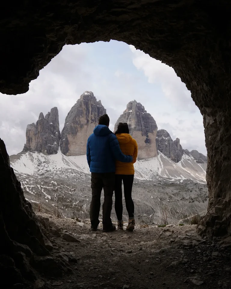 A couple sitting inside a mountain cave looking out at the Tre Cime di Lavaredo peaks.