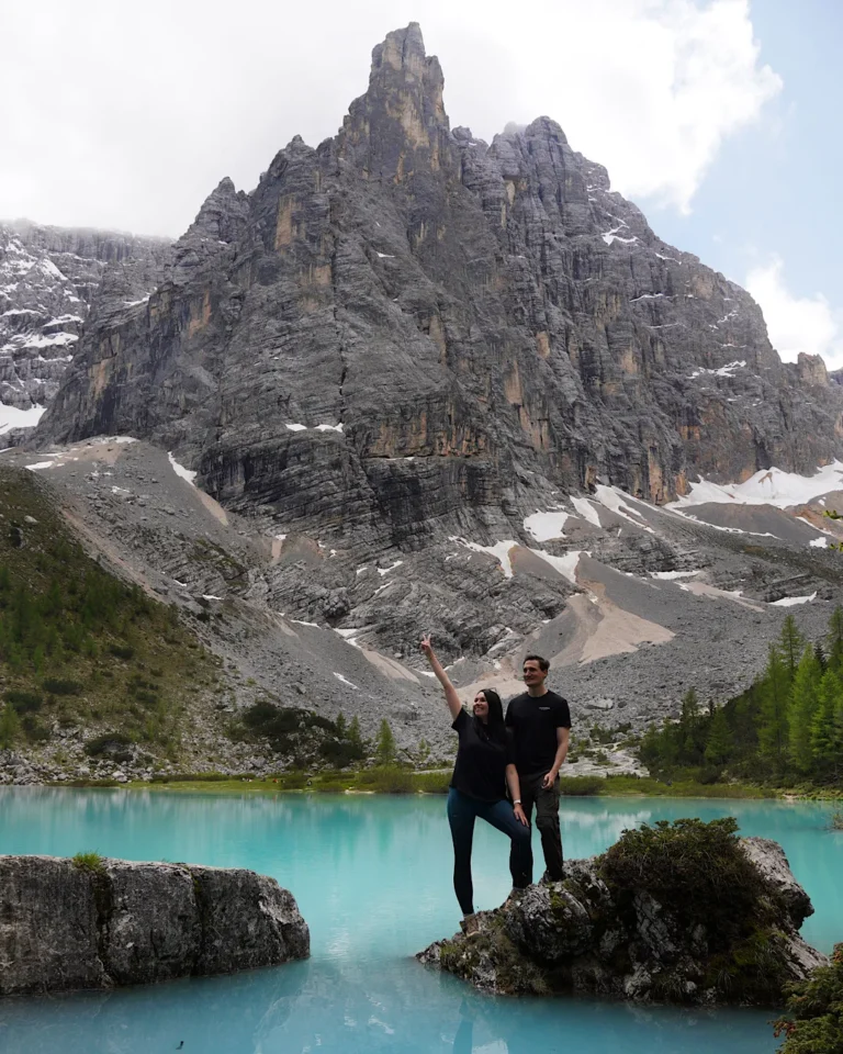 A couple standing on a rock in front of a bright turquoise mountain lake and towering cliffs in the Dolomites.