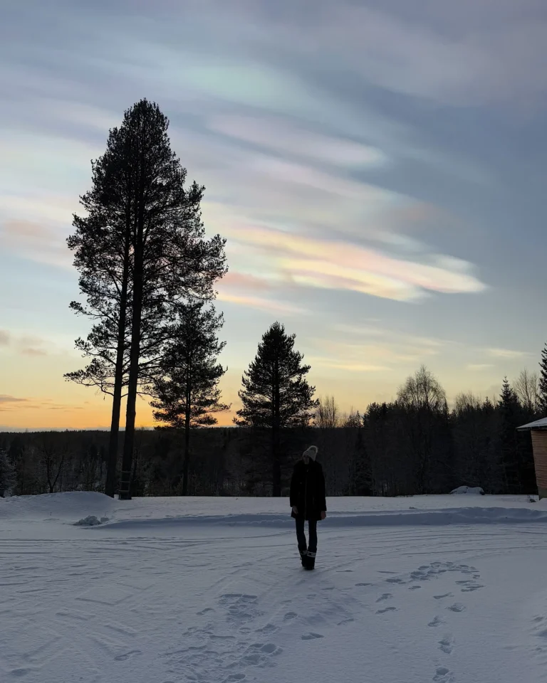 A woman standing in a snowy landscape in Lapland with rare, iridescent polar stratospheric clouds (nacreous clouds) glowing in the sky above her.