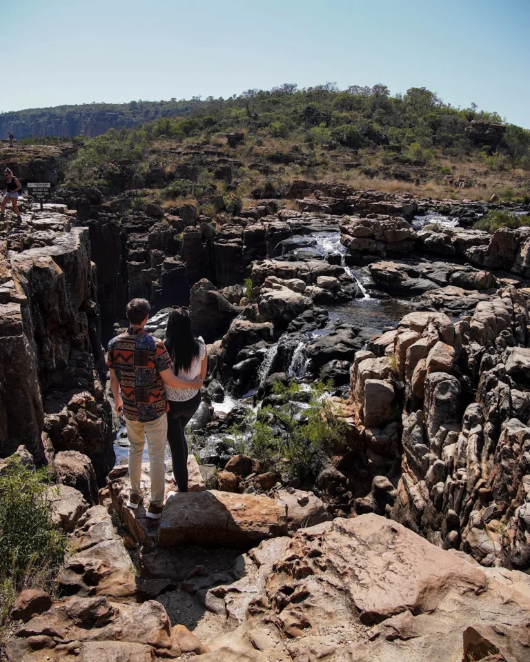 A travel couple standing with their backs to the camera at the top of Bourke's Luck Potholes, looking down.