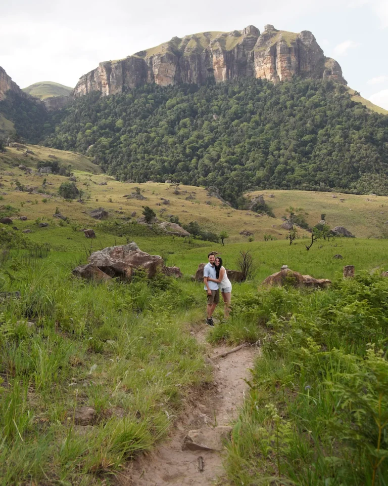 A happy travel couple hugging on a hiking trail in the Drakensberg in front of a mountain backdrop.