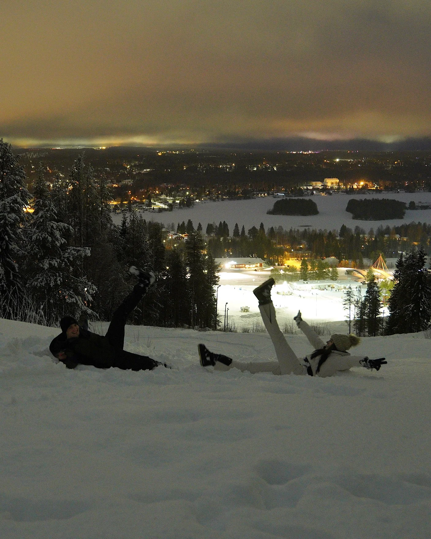 Ein Paar liegt abends im Schnee auf dem Berg Ounasvaara und blickt auf die Lichter von Rovaniemi.