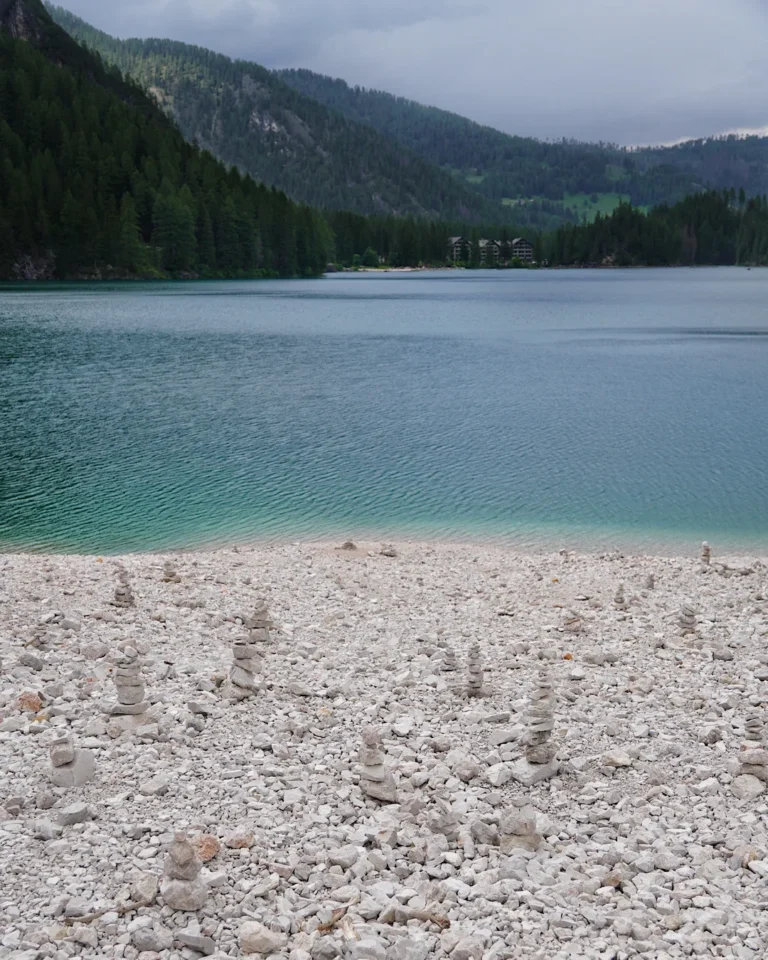 Zahlreiche kleine Steinmännchen auf einem weißen Kiesstrand am Ufer eines klaren Bergsees in den Alpen, Dolomiten.