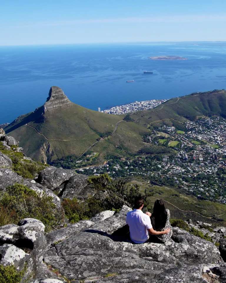 Travel couple sitting on Table Mountain looking down at Lions Head and the Cape Town skyline under a clear sky.