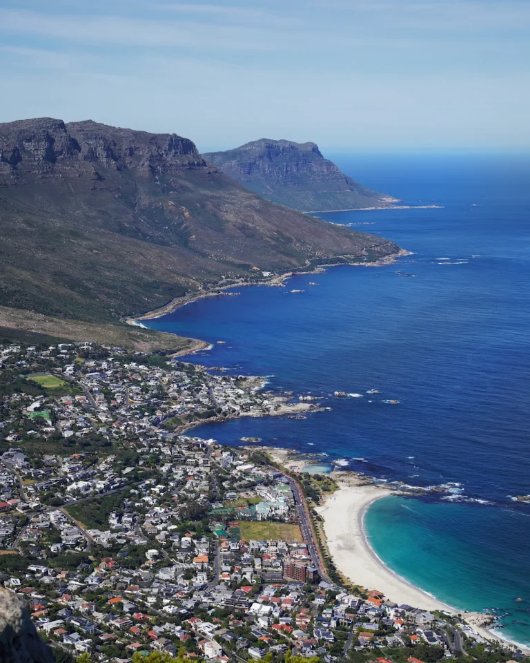Blick vom Tafelberg auf das dunkelblaue Meer, hellblaue Küstenstreifen und die Häuser der Stadt bei Sonnenschein.