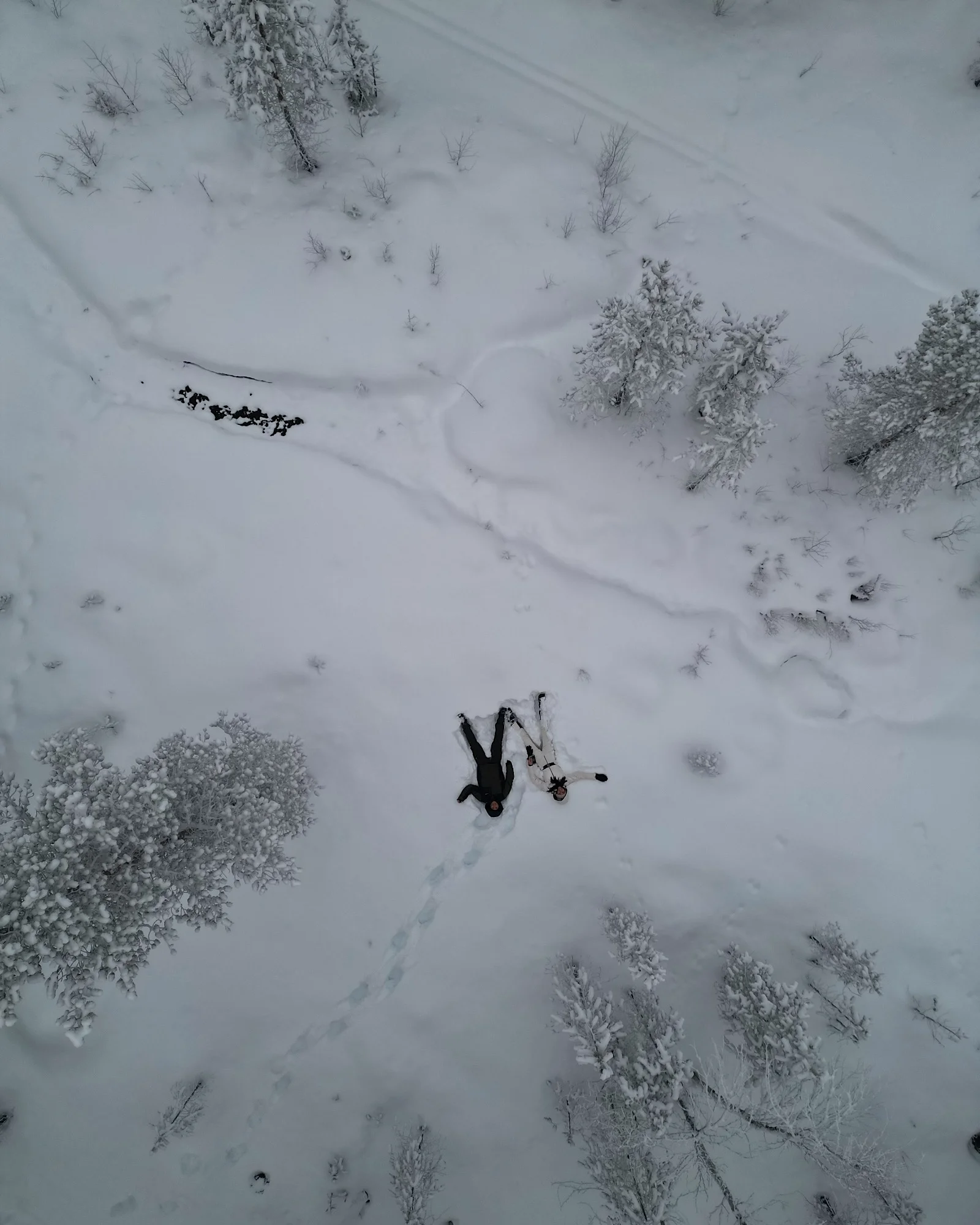 Draufsicht einer Drohne auf ein Paar, das im tiefen Schnee mitten in einem verschneiten Wald in Lappland liegt.