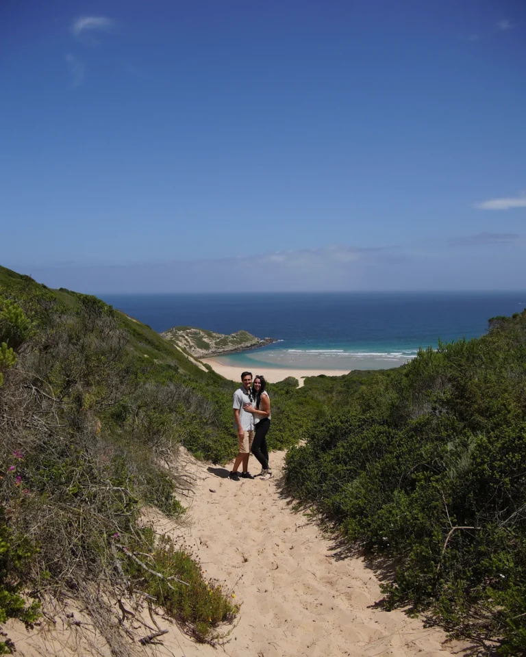 Junges Paar umarmt sich auf dem Wanderweg im Robberg Nature Reserve mit Blick auf die blaue Bucht.