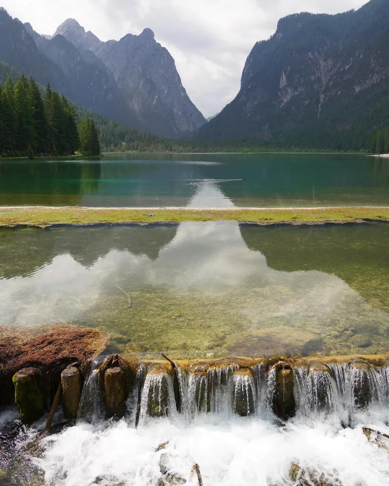 Ein kleiner künstlicher Wasserfall am Abfluss des Toblacher Sees mit Bergen im Hintergrund.