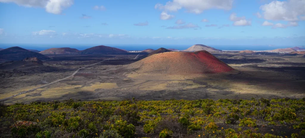 A panoramic photograph of Lanzarote's volcanic landscape during a hike, featuring a red-hued cinder cone volcano, dark lava fields, a winding trail, and the deep blue ocean under a partly cloudy sky.